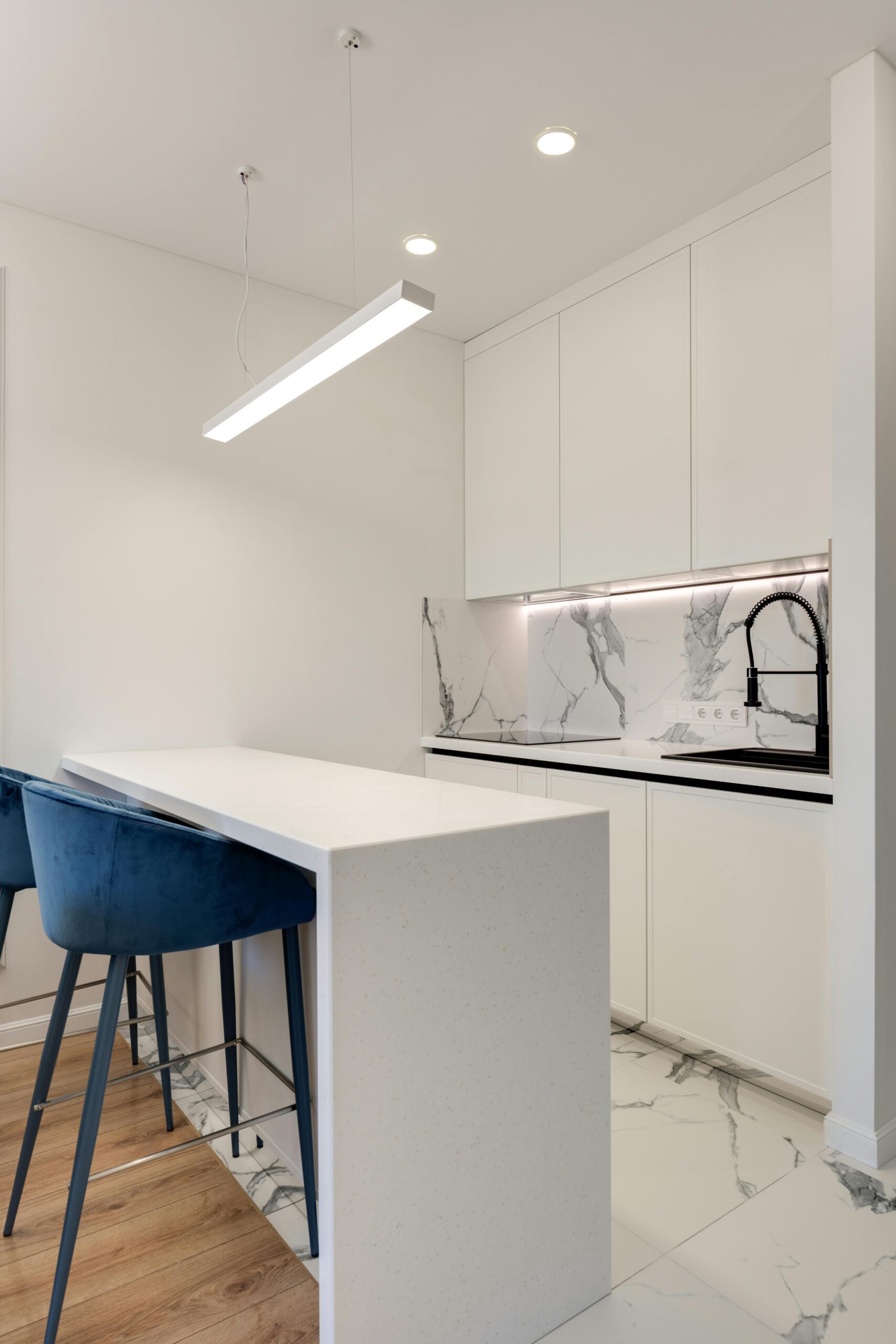 Compact kitchen featuring white quartz countertops and marble backsplash, with blue velvet bar stools for a pop of color and modern, sophisticated lighting.